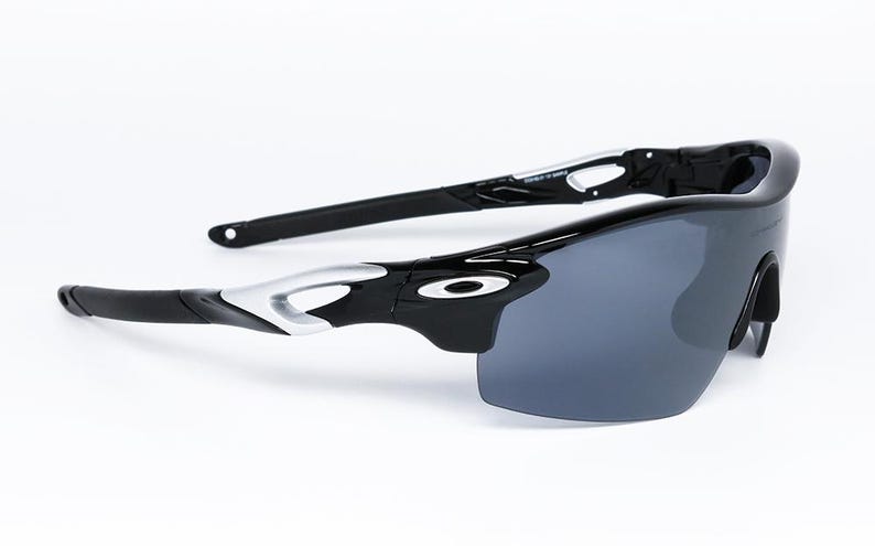 May include: Black and silver sports sunglasses with dark gray lenses. The sunglasses have a sleek, modern design with a curved frame and a prominent brand logo on the temples. The lenses are a dark gray color.