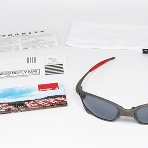 May include: Gray Oakley sunglasses with red accents on the temples, resting on a white surface. Included are a white pouch, paperwork, and a Ducati-branded card featuring a racetrack scene. The sunglasses have dark lenses.