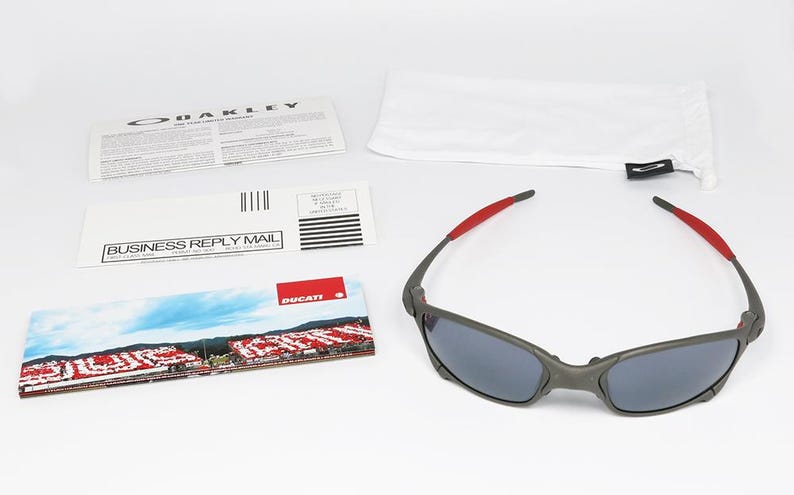 May include: Gray Oakley sunglasses with red accents on the temples, resting on a white surface. Included are a white pouch, paperwork, and a Ducati-branded card featuring a racetrack scene. The sunglasses have dark lenses.