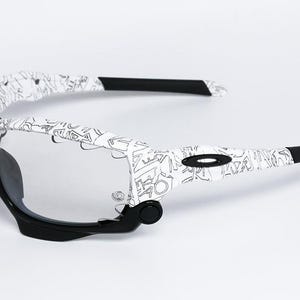 May include: White and black sports sunglasses with a unique pattern on the arms. The sunglasses have a black frame and clear lenses. The arms are white with a black graphic design. The Oakley logo is visible on the side.