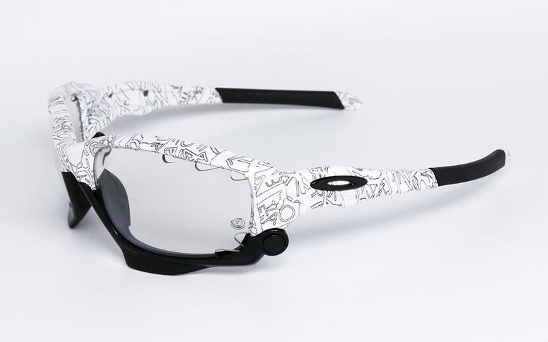 May include: White and black sports sunglasses with a unique pattern on the arms. The sunglasses have a black frame and clear lenses. The arms are white with a black graphic design. The Oakley logo is visible on the side.