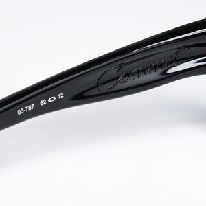 May include: Close-up of black sunglasses with the word "Commit" in cursive script on the arm. The arm also features the numbers "03-787 62 12". The lenses are a dark gray color.