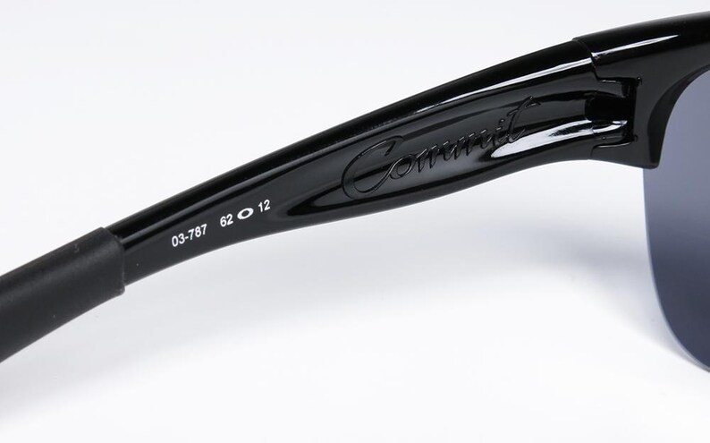 May include: Close-up of black sunglasses with the word "Commit" in cursive script on the arm. The arm also features the numbers "03-787 62 12". The lenses are a dark gray color.