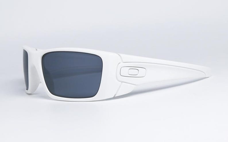 May include: White sunglasses with dark grey lenses. The sunglasses have a rectangular shape and a modern design. The frame is a solid white colour, and the lenses are a dark grey. The sunglasses are set against a white background.