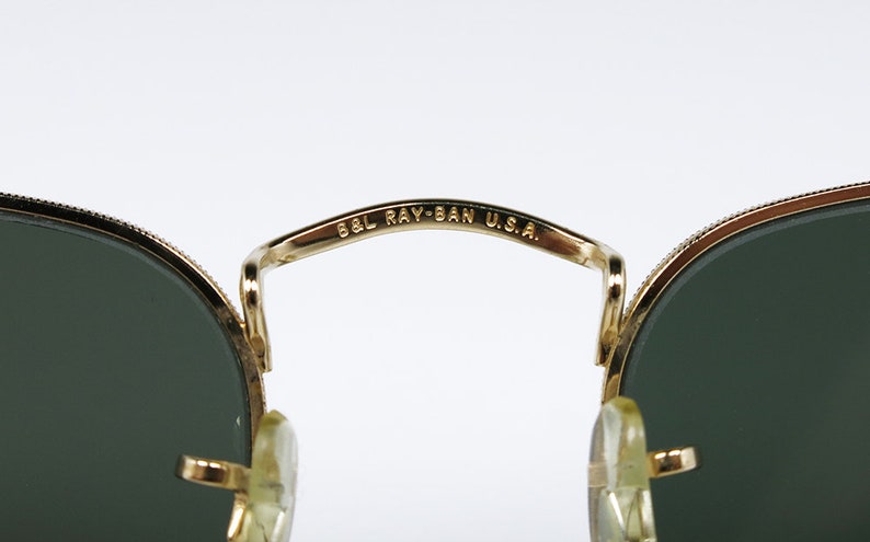 May include: Close-up of the gold-tone metal bridge of a pair of Ray-Ban sunglasses. The bridge is inscribed with "B&L RAY-BAN U.S.A."