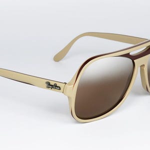 May include: Aviator-style sunglasses with a cream-colored frame and brown lenses. The sunglasses have a double bridge and the Ray-Ban logo is visible on the temple. The lenses have a gradient tint.