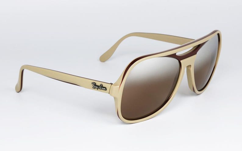 May include: Aviator-style sunglasses with a cream-colored frame and brown lenses. The sunglasses have a double bridge and the Ray-Ban logo is visible on the temple. The lenses have a gradient tint.