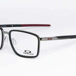 May include: Oakley Spindle eyeglasses with a black and silver frame and red temple tips. The glasses have a rectangular shape and the Oakley logo is visible on the lower right lens. The word "SPINDLE" is printed on the top of the frame.