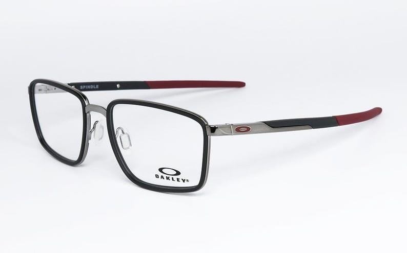 May include: Oakley Spindle eyeglasses with a black and silver frame and red temple tips. The glasses have a rectangular shape and the Oakley logo is visible on the lower right lens. The word "SPINDLE" is printed on the top of the frame.
