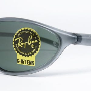 May include: Gray Ray-Ban sunglasses with dark green lenses. The sunglasses have a curved, wraparound design and the Ray-Ban logo is visible on the temple and lens. A yellow sticker on the lens reads "Ray-Ban Sunglasses by Luxottica G-15 Lens."
