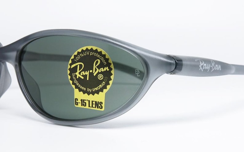 May include: Gray Ray-Ban sunglasses with dark green lenses. The sunglasses have a curved, wraparound design and the Ray-Ban logo is visible on the temple and lens. A yellow sticker on the lens reads "Ray-Ban Sunglasses by Luxottica G-15 Lens."