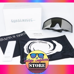 Puede incluir: Un par de gafas de sol negras y plateadas en una bolsa protectora con un logotipo blanco. Las gafas de sol están en una caja blanca con el texto "SUNGLASSES" y "MYKITA & BERM" impreso en ella.