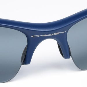 以下が含まれることがあります： グレーレンズの青いサングラスのクローズアップ。ブランド名「OAKLEY」がフレームにシルバーで表示されています。サングラスは、アウトドア活動に適した、洗練されたモダンなデザインです。