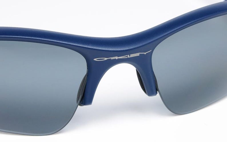 以下が含まれることがあります： グレーレンズの青いサングラスのクローズアップ。ブランド名「OAKLEY」がフレームにシルバーで表示されています。サングラスは、アウトドア活動に適した、洗練されたモダンなデザインです。