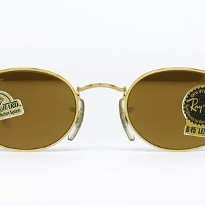 Pu&ograve; includere: Occhiali da sole ovali con montatura dorata e lenti marroni. Gli occhiali hanno un ponte e naselli. Gli adesivi sulle lenti recitano "DIAMOND HARD Scratch-Protection System" e "Ray-Ban B-15 LENS 100% UV protection".