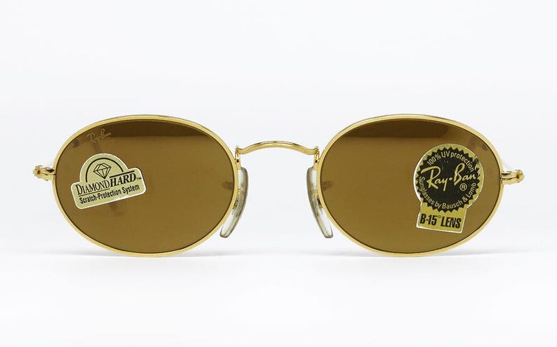 Pu&ograve; includere: Occhiali da sole ovali con montatura dorata e lenti marroni. Gli occhiali hanno un ponte e naselli. Gli adesivi sulle lenti recitano "DIAMOND HARD Scratch-Protection System" e "Ray-Ban B-15 LENS 100% UV protection".