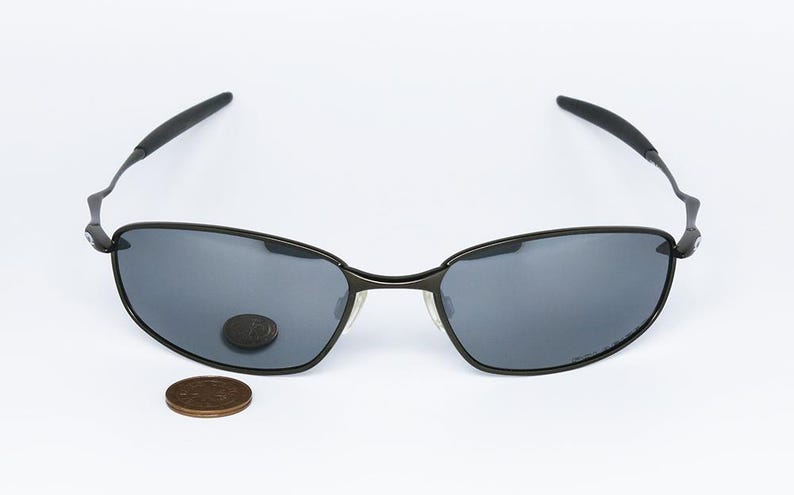 Może przedstawiać: Czarne okulary przeciwsłoneczne z ciemnoszarymi soczewkami, o eleganckim, nowoczesnym designie. Oprawki są czarne z cienkim metalowym mostkiem i zausznikami. Moneta leży w pobliżu okular&oacute;w.