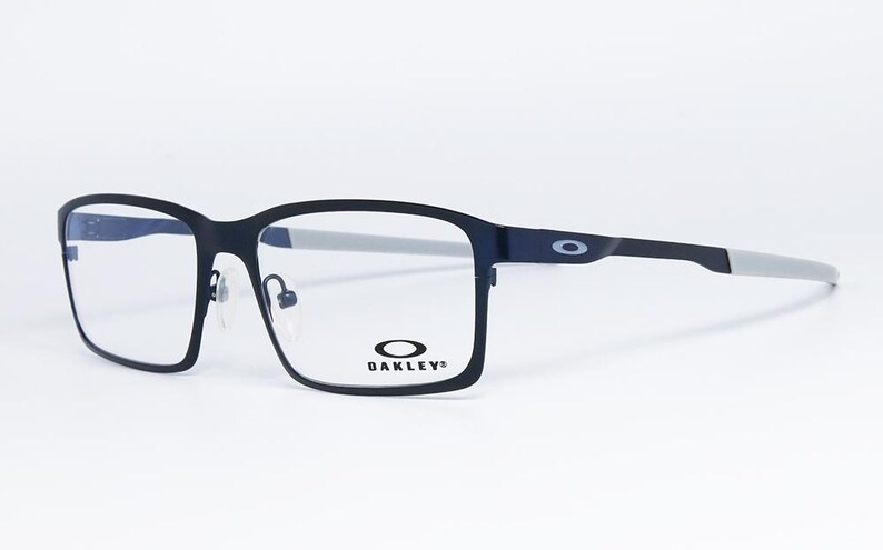 K&ouml;nnte beinhalten: Schwarze rechteckige Brille mit grauen B&uuml;geln. Die Brille hat eine matte Oberfl&auml;che und das Oakley-Logo ist auf dem Rahmen und dem B&uuml;gel sichtbar. Die Gl&auml;ser sind klar.