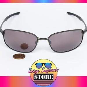 May include: Dark grey sunglasses with tinted lenses. A coin rests on one lens and another below. The glasses are on a white surface. The logo "Vintage Sunglasses Store" is at the bottom.