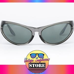 May include: Gray sunglasses with dark lenses. The sunglasses have a curved frame and a subtle logo on the temple. The image includes a logo for a vintage sunglasses store.