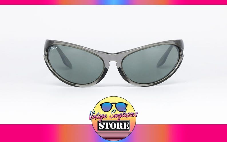 May include: Gray sunglasses with dark lenses. The sunglasses have a curved frame and a subtle logo on the temple. The image includes a logo for a vintage sunglasses store.