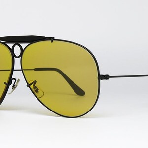 Puede incluir: Gafas de sol estilo aviador con montura negra y lentes amarillas. Las gafas tienen un doble puente y una barra superior. Las patillas son rectas con extremos curvados. Las lentes son de color amarillo brillante.