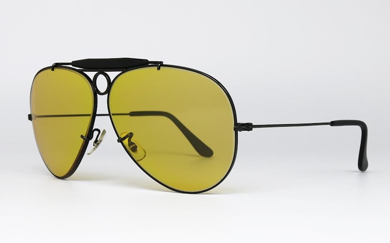 Puede incluir: Gafas de sol estilo aviador con montura negra y lentes amarillas. Las gafas tienen un doble puente y una barra superior. Las patillas son rectas con extremos curvados. Las lentes son de color amarillo brillante.