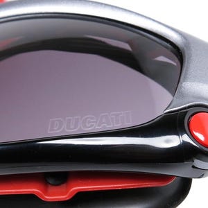 Puede incluir: Primer plano de unas gafas de sol negras y plateadas con un bot&oacute;n rojo en el lateral. Las gafas de sol tienen una lente gris oscura con la palabra "DUCATI" grabada en la lente.