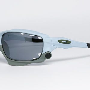 May include: Light blue and gray sports sunglasses with dark lenses. The sunglasses have a modern design with a gray frame and black earpieces. The Oakley logo is visible on the temple.
