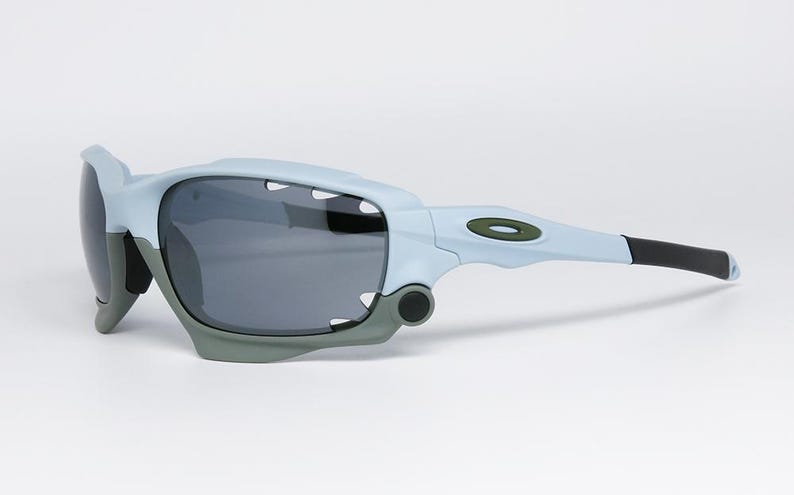 May include: Light blue and gray sports sunglasses with dark lenses. The sunglasses have a modern design with a gray frame and black earpieces. The Oakley logo is visible on the temple.