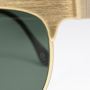 Puede incluir: Primer plano de gafas de sol doradas con lentes verdes. La montura tiene un acabado texturizado. Las gafas de sol tienen un peque&ntilde;o logotipo grabado en la lente.