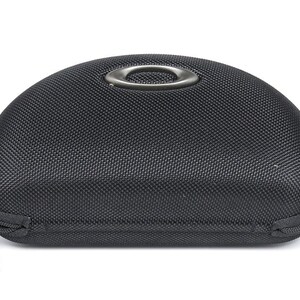 May include: Black, semi-hard case with a textured surface and a zipper closure. The case has a metal ring on top and a zipper pull with the Oakley logo. The case is designed to protect eyewear.