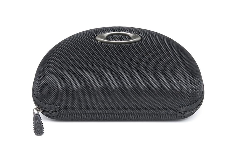 May include: Black, semi-hard case with a textured surface and a zipper closure. The case has a metal ring on top and a zipper pull with the Oakley logo. The case is designed to protect eyewear.