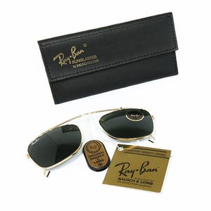 May include: Gold-framed Ray-Ban sunglasses with dark lenses, a black leather case, and a small brown case. The Ray-Ban logo is visible on the sunglasses and case. A tag with the Ray-Ban logo is also present.