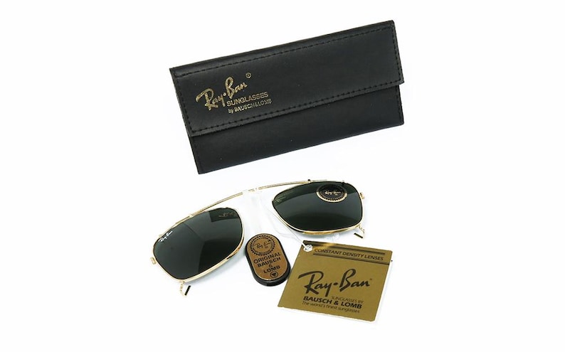 May include: Gold-framed Ray-Ban sunglasses with dark lenses, a black leather case, and a small brown case. The Ray-Ban logo is visible on the sunglasses and case. A tag with the Ray-Ban logo is also present.