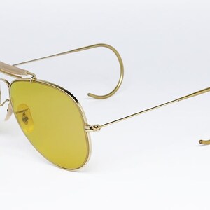 K&ouml;nnte beinhalten: Goldene Aviator-Sonnenbrille mit gelben Gl&auml;sern und einem Drahtgestell. Die Sonnenbrille hat ein einzigartiges Design mit einem gebogenen Drahtb&uuml;gel.
