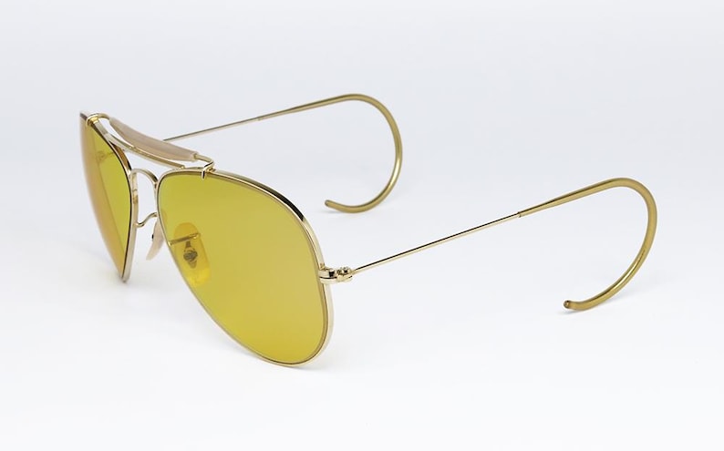 K&ouml;nnte beinhalten: Goldene Aviator-Sonnenbrille mit gelben Gl&auml;sern und einem Drahtgestell. Die Sonnenbrille hat ein einzigartiges Design mit einem gebogenen Drahtb&uuml;gel.