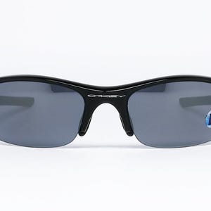 K&ouml;nnte beinhalten: Schwarze Oakley-Sonnenbrille mit grauen Gl&auml;sern. Die Sonnenbrille hat ein halb rahmenloses Design und einen geschwungenen Rahmen. Das Oakley-Logo ist auf der Br&uuml;cke sichtbar. Auf dem rechten Glas befindet sich ein blau-wei&szlig;er Aufkleber.