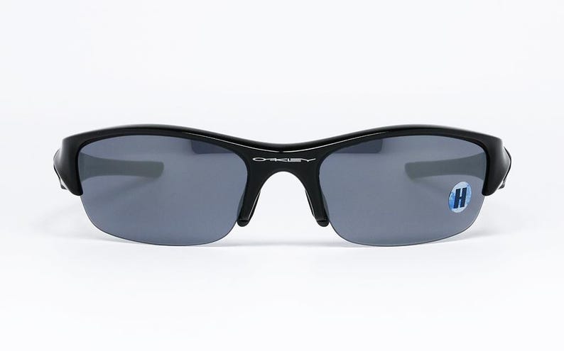 K&ouml;nnte beinhalten: Schwarze Oakley-Sonnenbrille mit grauen Gl&auml;sern. Die Sonnenbrille hat ein halb rahmenloses Design und einen geschwungenen Rahmen. Das Oakley-Logo ist auf der Br&uuml;cke sichtbar. Auf dem rechten Glas befindet sich ein blau-wei&szlig;er Aufkleber.