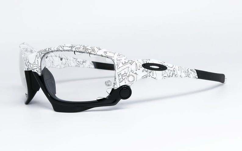 May include: White and black patterned sunglasses with clear lenses. The frames feature a unique design with black accents on the arms and nose bridge. The sunglasses are designed for sports and outdoor activities.