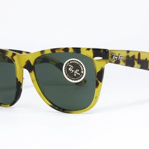 May include: Tortoiseshell-patterned sunglasses with dark lenses. The frames are yellow and black, with the Ray-Ban logo visible on the temple and lens. A round sticker with the Ray-Ban logo is on the lens.