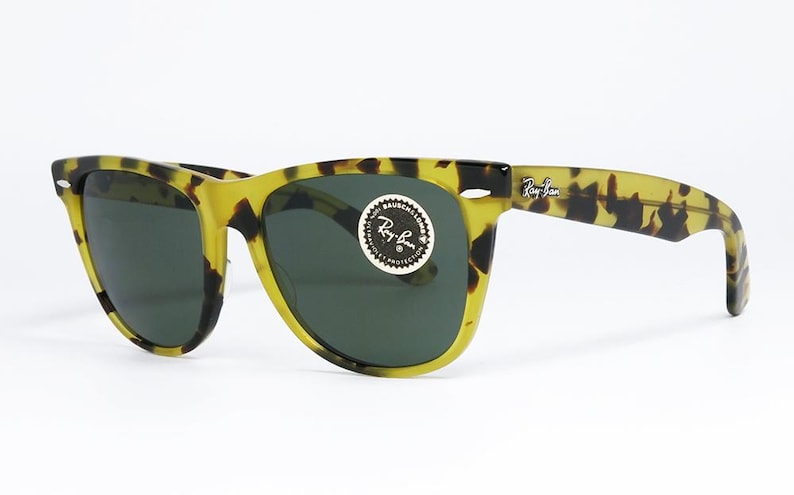 May include: Tortoiseshell-patterned sunglasses with dark lenses. The frames are yellow and black, with the Ray-Ban logo visible on the temple and lens. A round sticker with the Ray-Ban logo is on the lens.