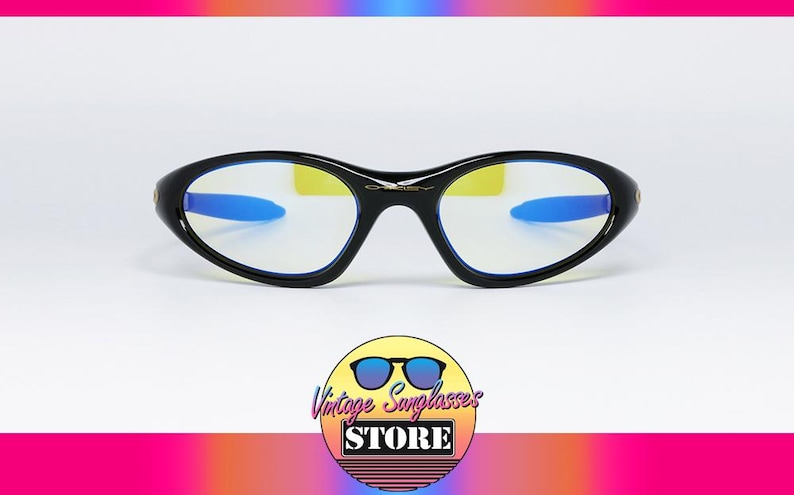 Peut inclure: Lunettes de soleil Oakley noires avec une monture incurv&eacute;e et des accents bleus sur les branches. Les verres ont une teinte jaune. Les lunettes de soleil sont affich&eacute;es sur un fond blanc. Le logo de Vintage Sunglasses Store est en bas.