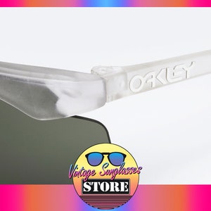 Puede incluir: Primer plano de una montura de gafas de sol Oakley blanca con el logotipo de Oakley en relieve en el lateral. Las gafas tienen lentes grises oscuras.