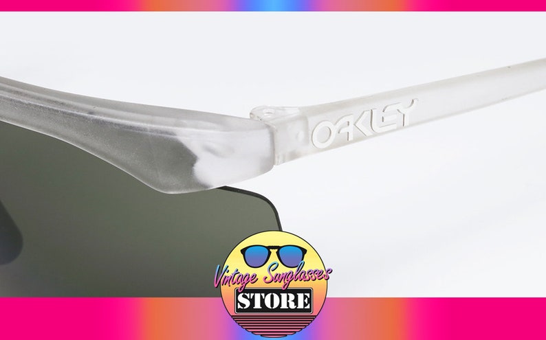 Puede incluir: Primer plano de una montura de gafas de sol Oakley blanca con el logotipo de Oakley en relieve en el lateral. Las gafas tienen lentes grises oscuras.
