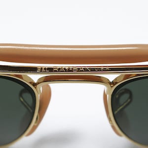 May include: Close-up of gold-framed sunglasses with dark green lenses. The top bar features a tan-colored bridge. The text "B&L RAY-BAN U.S.A." is visible on the frame. Classic eyewear.