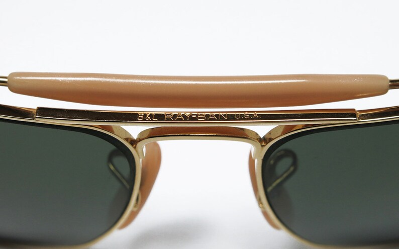 May include: Close-up of gold-framed sunglasses with dark green lenses. The top bar features a tan-colored bridge. The text "B&L RAY-BAN U.S.A." is visible on the frame. Classic eyewear.