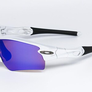 Pu&ograve; includere: Occhiali da sole sportivi bianchi e argento con lenti a specchio viola. Gli occhiali hanno terminali neri e un design elegante. Il logo Oakley &egrave; visibile.