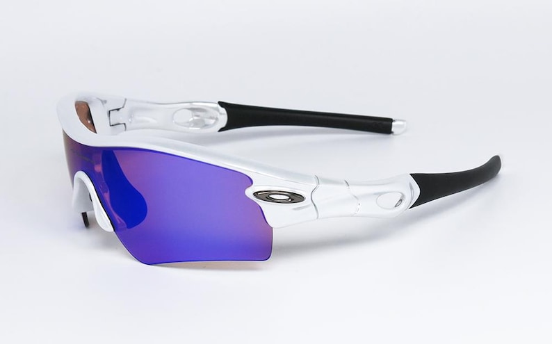 Pu&ograve; includere: Occhiali da sole sportivi bianchi e argento con lenti a specchio viola. Gli occhiali hanno terminali neri e un design elegante. Il logo Oakley &egrave; visibile.