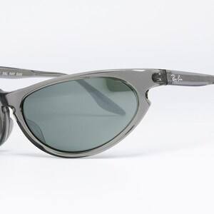 May include: Gray Ray-Ban sunglasses with a wraparound design. The lenses are a dark green color, and the frames are a translucent gray. The Ray-Ban logo is visible on the temple.